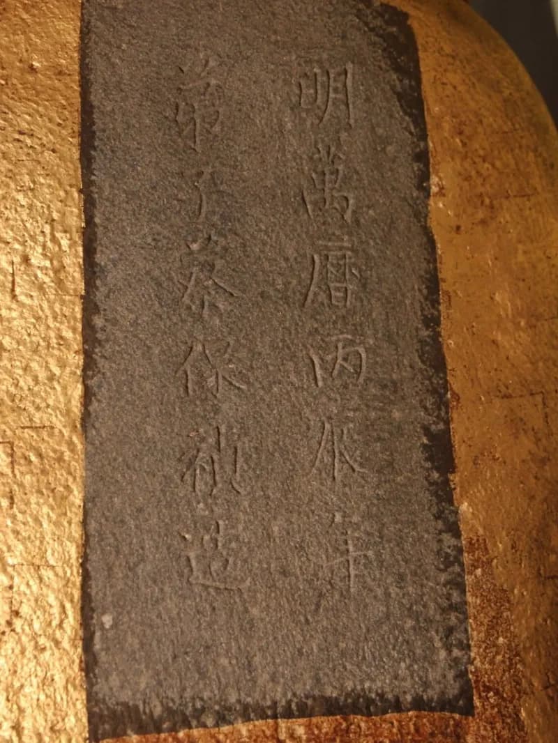 鎮殿觀音佛祖石像背面刻款特寫：金色石像背上一塊深色刻面，兩行陰刻，右行「明萬曆丙辰年」，左行為造像弟子題名