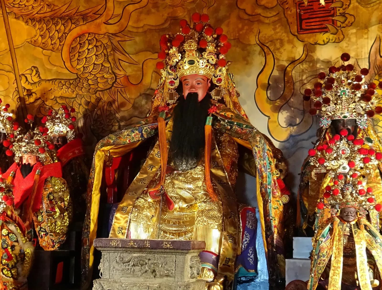 前殿主祀保生大帝神像半側面：頭戴紅絨球帝冠、黑色長鬚，身披金色神袍，背後是金色龍紋壁畫（攝於 2015 年）